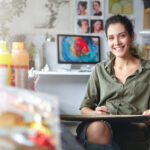 smiling female artist wearing casual clothes sitting her cabinet with sketches colorful paint having happy expression while being glad create beautiful picture painter working workshop